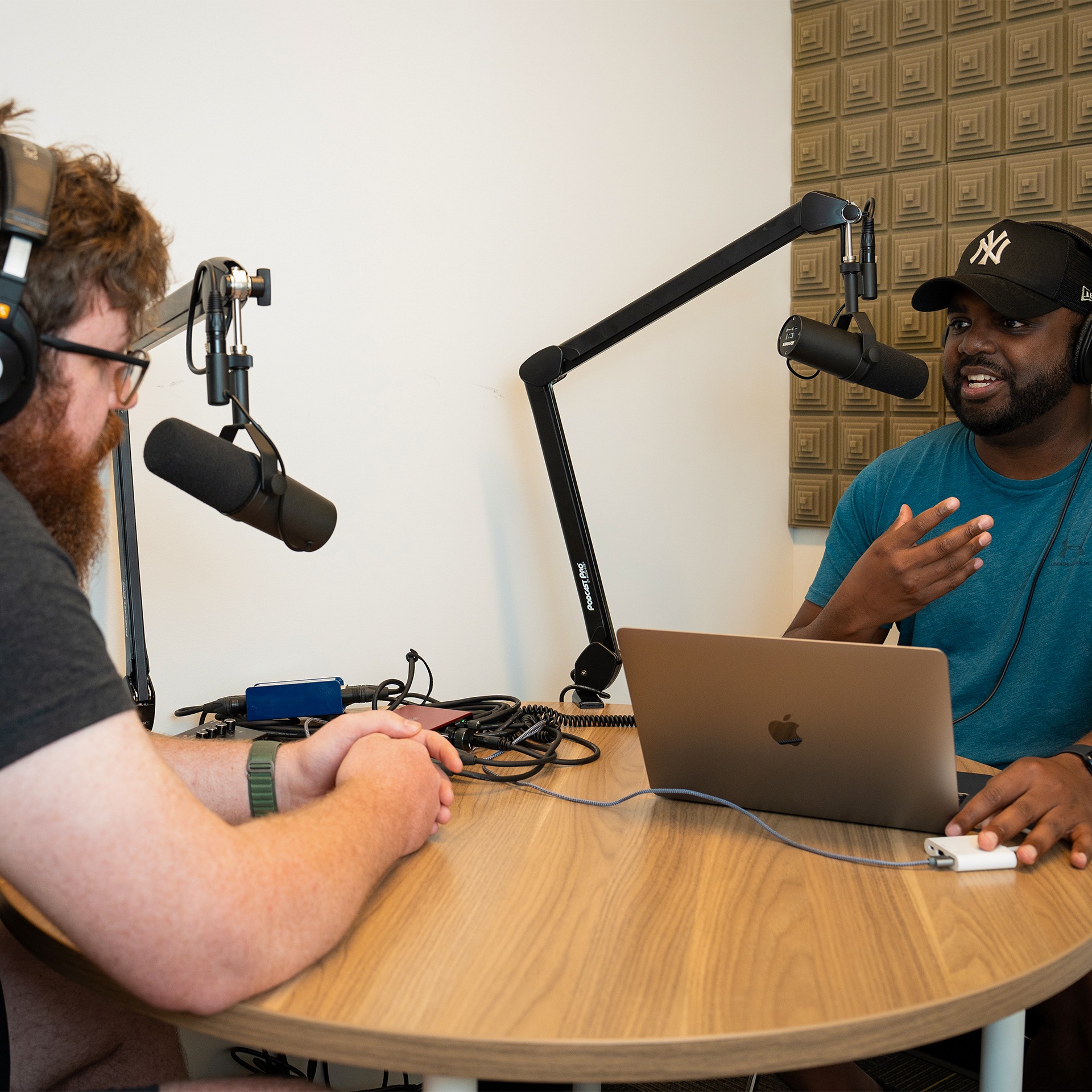 Two members of The Factory sitting at a round wooden table with microphones in front of their mouths pretending to have a discussion.