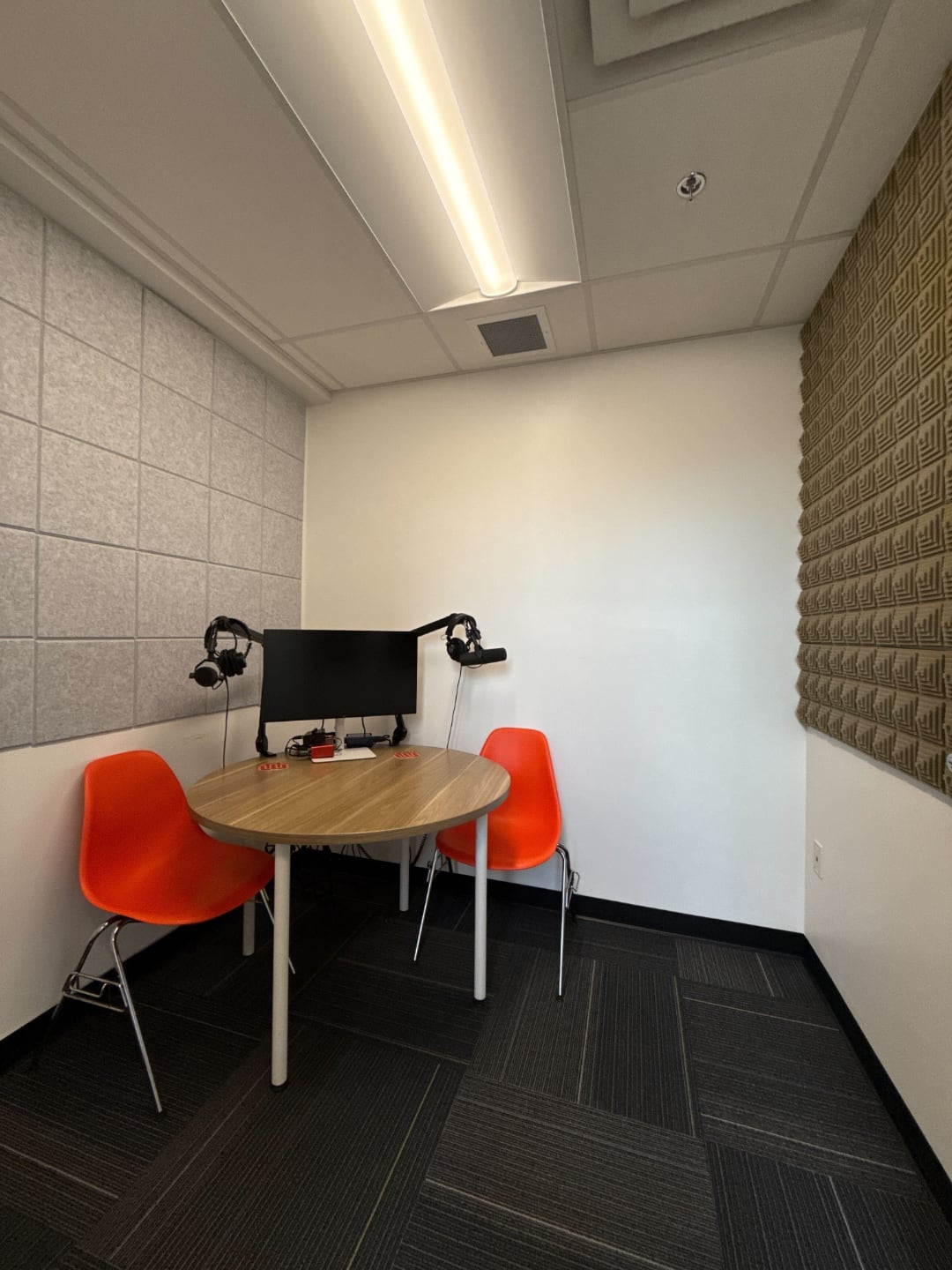 Photo of The Factory podcast booth with two orange plastic chairs and an a wooden round desk placed in the corner of the west side of the room