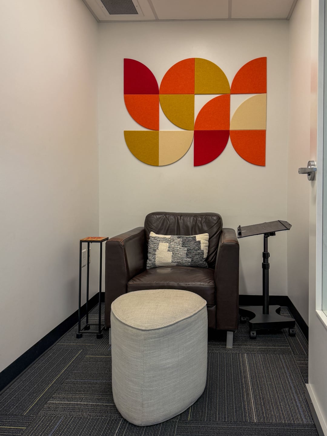 Photo of The Factory privacy and maternity booth with an large brown leather chair and light gray colored foot stool a black laptop stand and a wooden drinking cup desk